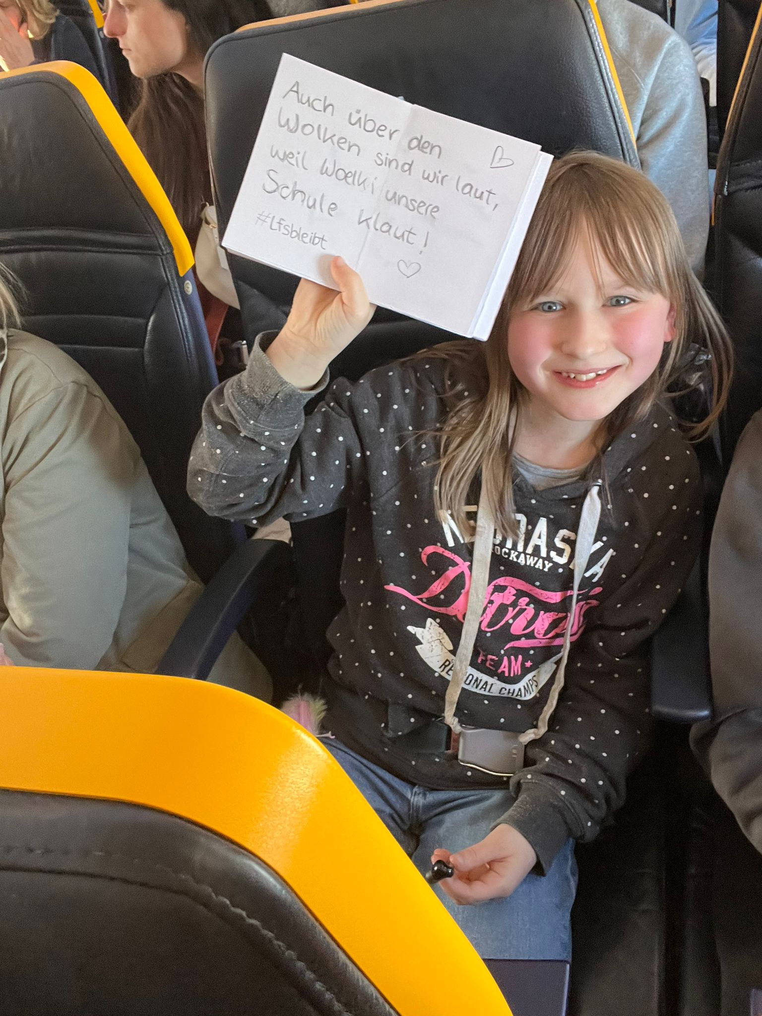 Mädchen mit Plakat "Auch über den Wolken sind wir laut, weil Woelki unsere Schule klaut!" im Flugzeug.