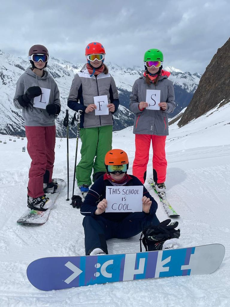 Skifahrer mit "L", "F", "S" und "This School is cool" Plakaten im Schnee.