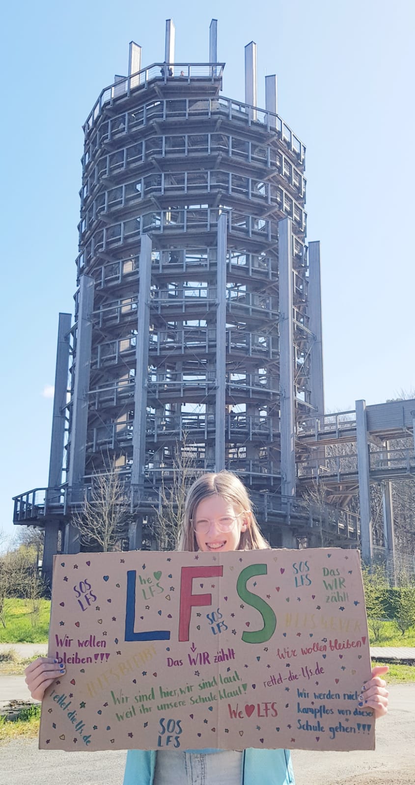 Eine Schülerin steht vor dem Panarbora Aussichtsturm mit einem LFS Plakat